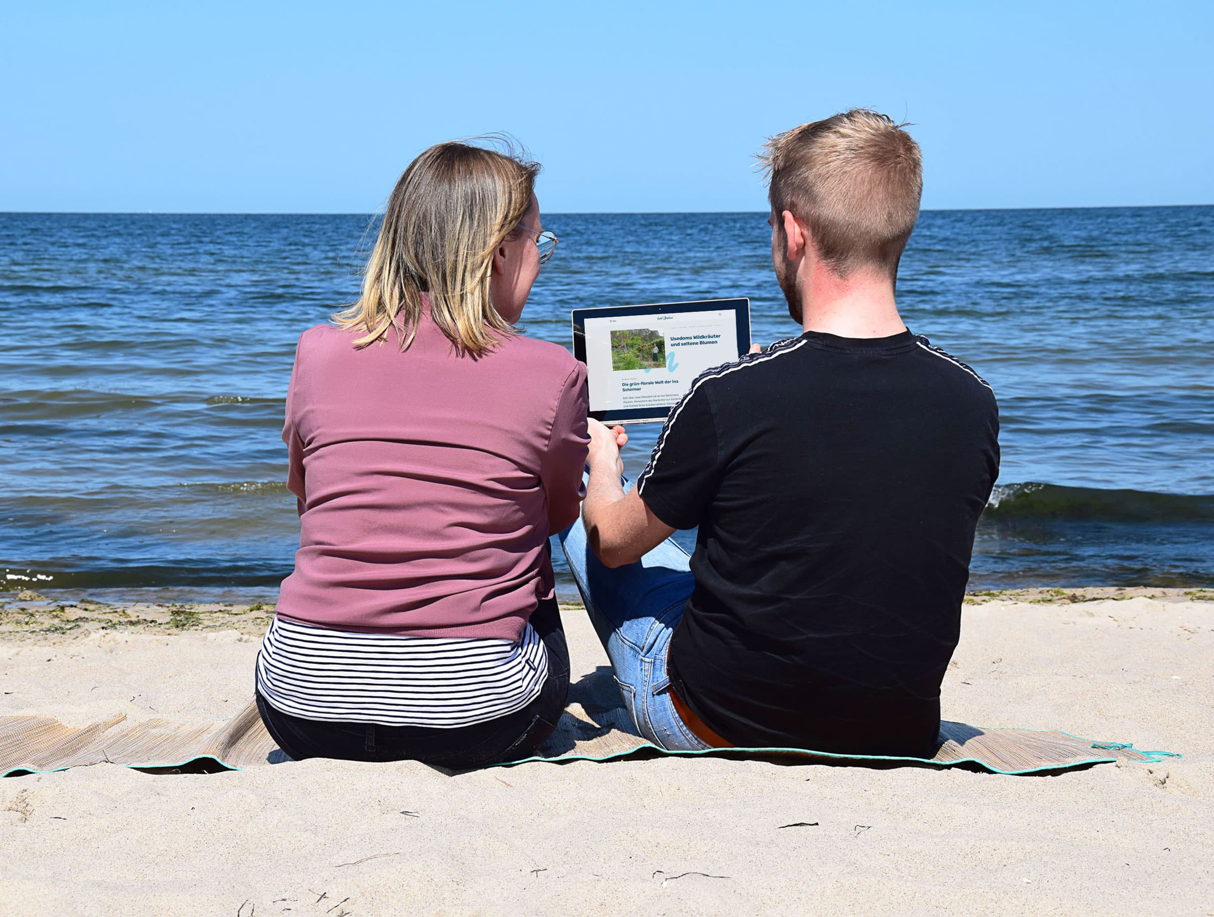 Pärchen sitzt am Strand und liest auf einem Tablet.